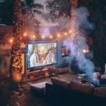 A night time image of a small cinema setup in a backyard with furniture set out, fairy light hung and trees surrounding